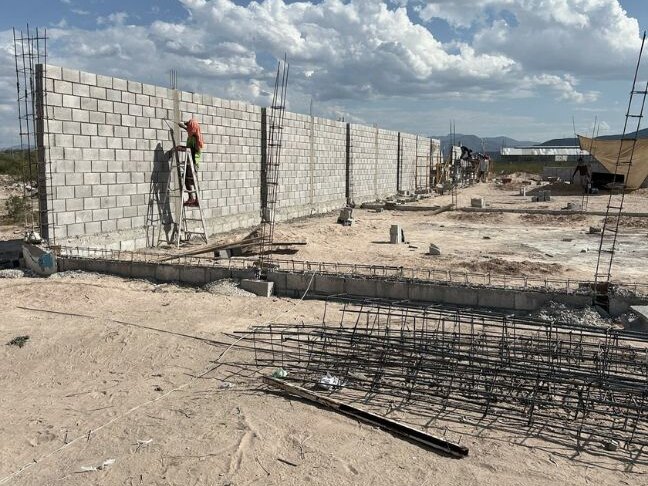 Muro de block de la bodega en obra, con castillos de varilla y un trabajador sobre una escalera; en primer plano, armados de acero sobre la arena.